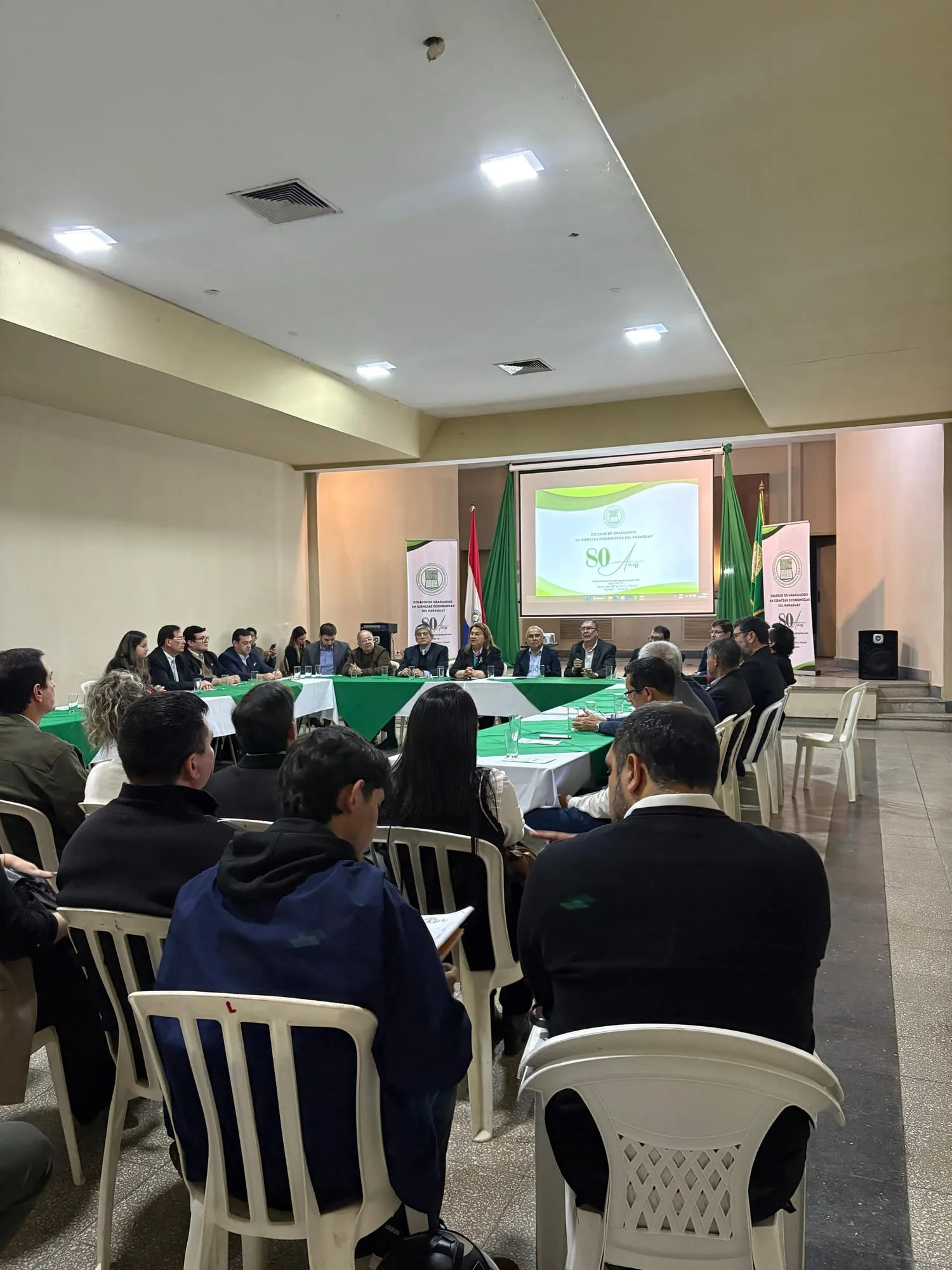 Vista general del salón, con la mesa directiva al fondo y la pantalla del 80 aniversario del Colegio